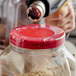 A person using a drill to shrink a clear band around a jar with a red lid.
