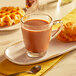 A glass cup of Swiss Miss hot chocolate on a plate with waffles and a spoon.