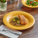 A yellow Acopa Foundations wide rim melamine plate with a piece of fish and vegetables on a table.