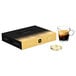 A black Nespresso box with gold and black lettering on it on a table with a glass of coffee with a gold lid.