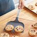 A person using a Wilton "The Really Big Spatula" to lift a cookie with chocolate chunks off a tray.