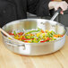 A person sautéing vegetables in a Vollrath Wear-Ever saute pan on a table.
