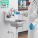 A person in a blue lab coat and gloves washing hands in a Regency wall mounted sink.