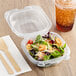 A salad in a Genpak clear plastic hinged container with a wooden fork next to a drink.