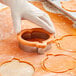 A gloved hand using a Wilton metal pumpkin cookie cutter to cut out a cookie.