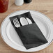 A fork and knife on a Hoffmaster black square dinner napkin on a white plate.