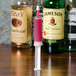 A Jell-O syringe filled with pink liquid on a counter next to bottles of liquor.