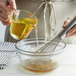 A person pouring yellow corn oil from a measuring cup into a bowl with a whisk.