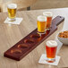 An Acopa mahogany beer flight paddle with glasses of beer and a bowl of pretzels on a table.