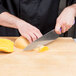 A person's hand using a Mercer Culinary purple chef knife to cut a squash on a wooden cutting board.