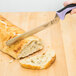 A hand holding a Mercer Culinary purple bread knife cutting a piece of bread on a cutting board.