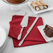 A table with a cake on a glass stand and the American Metalcraft hammered stainless steel serrated knife on a red napkin.