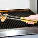 A person holding a pair of Vollrath stainless steel scalloped tongs with yellow Kool Touch handles over a grill.