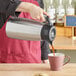 A person using a Curtis stainless steel coffee server to pour coffee into a cup.