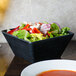 A black square melamine bowl of salad on a table next to a bowl of soup.