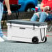 A person wheeling a white CaterGator outdoor cooler with black wheels.