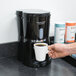 A person holding a white mug of coffee in front of a Hamilton Beach BrewStation coffee maker.
