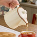 A cream-colored porcelain teapot with a lid being used to pour tea into a cup.