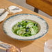 A round gray porcelain wide rim bowl filled with a green salad, cucumber slices, blueberries, and cheese crumbles.