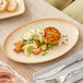 An oval beige porcelain platter displaying a meal with potatoes, lemon slices, cucumber, and a crab cake.