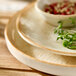 A round beige porcelain plate with a slightly rustic edge, shown stacked with other plates and a small bowl.