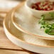 A round beige porcelain plate with a slightly rustic edge, shown stacked with other plates and a small bowl.