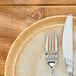 A round beige porcelain plate with a subtle speckled pattern, shown with a fork and knife on top.