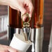 A person pouring liquid into a cup from a Vollrath brass beverage dispenser.