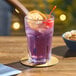 A Acopa Memphis highball glass filled with purple liquid, a cherry, and an orange slice on a table with a bowl of popcorn.
