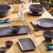 A set of dark gray round melamine plates and bowls arranged on a wooden dining table with placemats and utensils.