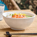 A white round melamine bowl filled with food, placed on a bamboo mat with chopsticks and a glass of water nearby.