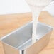 A spoon pouring white liquid into a Vollrath stainless steel bread loaf pan.
