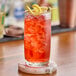 A clear convex 14 oz. beverage glass filled with a red iced drink and garnished with a lemon twist and olive on a skewer.
