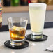 A clear Acopa Fusion 15.25 oz. beverage glass filled with a light-colored drink, shown next to a smaller glass with an amber liquid and ice, both on black coasters.