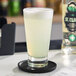 A clear Acopa Fusion beverage glass filled with a light yellow drink and topped with foam, placed on a black coaster.