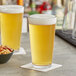 A clear 20 oz. mixing glass filled with light beer, shown on a bar counter with a bowl of mixed nuts nearby.