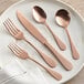 A five-piece set of rose gold stainless steel flatware including two forks, a knife, and two spoons displayed on a white plate.