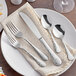 A five-piece set of stainless steel flatware with a textured handle design, including two forks, a knife, and two spoons, displayed on a napkin and plate.
