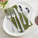 A five-piece set of stainless steel flatware including two forks, a knife, and two spoons, displayed on a green napkin and white plates.