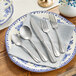 A five-piece set of distressed stainless steel flatware including two forks, two spoons, and a knife, displayed on a plate with a napkin.