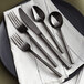 A five-piece set of black stainless steel flatware including two forks, a knife, and two spoons, displayed on a white napkin and dark plate.