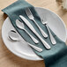 A five-piece set of stainless steel flatware including two forks, two spoons, and a knife, displayed on a white plate with a teal napkin.