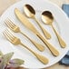A five-piece set of gold-colored stainless steel flatware, including two forks, a knife, and two spoons, displayed on a white plate.