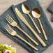 A five-piece set of gold-colored stainless steel flatware, including two forks, a knife, and two spoons, displayed on a dark cloth napkin.