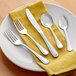 A five-piece set of stainless steel flatware including two forks, a knife, and two spoons, displayed on a yellow napkin and white plate.