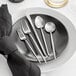 A five-piece set of brushed stainless steel flatware, including two forks, a knife, and two spoons, displayed on a black plate with a white plate and black napkin.