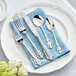 A five-piece set of ornate stainless steel flatware, including two forks, a knife, and two spoons, displayed on a blue napkin atop a white plate.
