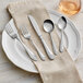 A five-piece set of stainless steel flatware with decorative handles, displayed on a white plate with a beige napkin.