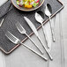 A five-piece set of stainless steel flatware including two forks, a knife, and two spoons, displayed on a table setting.