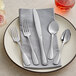 A five-piece set of stainless steel flatware including two forks, a knife, and two spoons, arranged on a plate with a gray napkin.